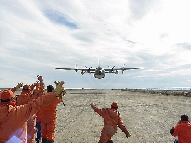 Varias personas con uniforme naranja levantan las manos para recibir a un avión Hércules que está pronto a aterrizar.