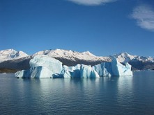 Bloque de hielo rodeado de agua muy azul. De fondo, montañas nevadas.