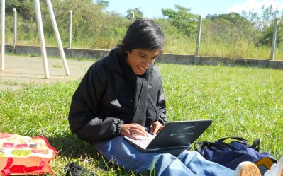 Adolescente sentado en el pasto, al aire libre, con netbook. Sonríe mirando la pantalla