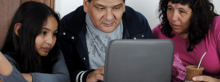 Un papá, una mamá y su hija pequeña observan atentos la pantalla de una netbook, en su casa. En color.