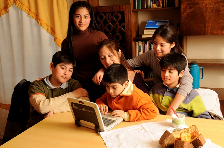 Madre rodeada por su cinco hijos e hijas: tres varones y dos mujeres. La madre está sentada, junto a sus tres hijos varones: el más pequeño manipula una netbook. Las dos hijas están paradas. Todos están en la sala comedor de su casa. En color.