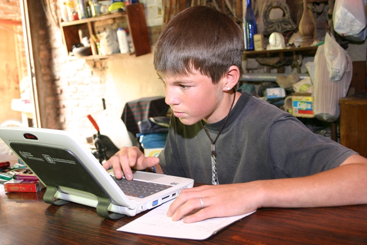 Alumno de escuela rural con su netbook, sentado en un ambiente de su casa. El adolescente mira fijamente la pantalla. En color.
